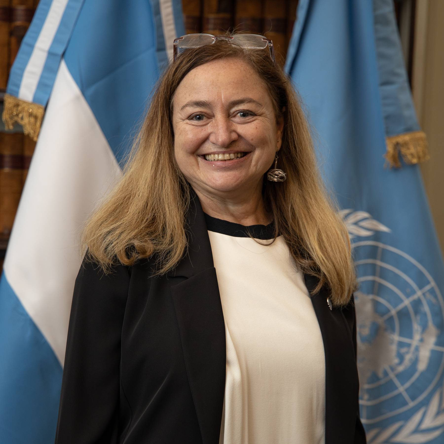 A woman in a black jacket and white shirt smiles brightly at the camera next to the United Nations and Argentinian flags.