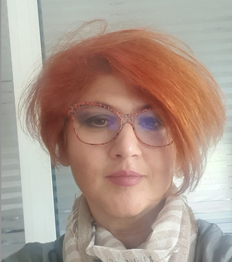 A woman with red hair and peach glasses looks directly at the camera.