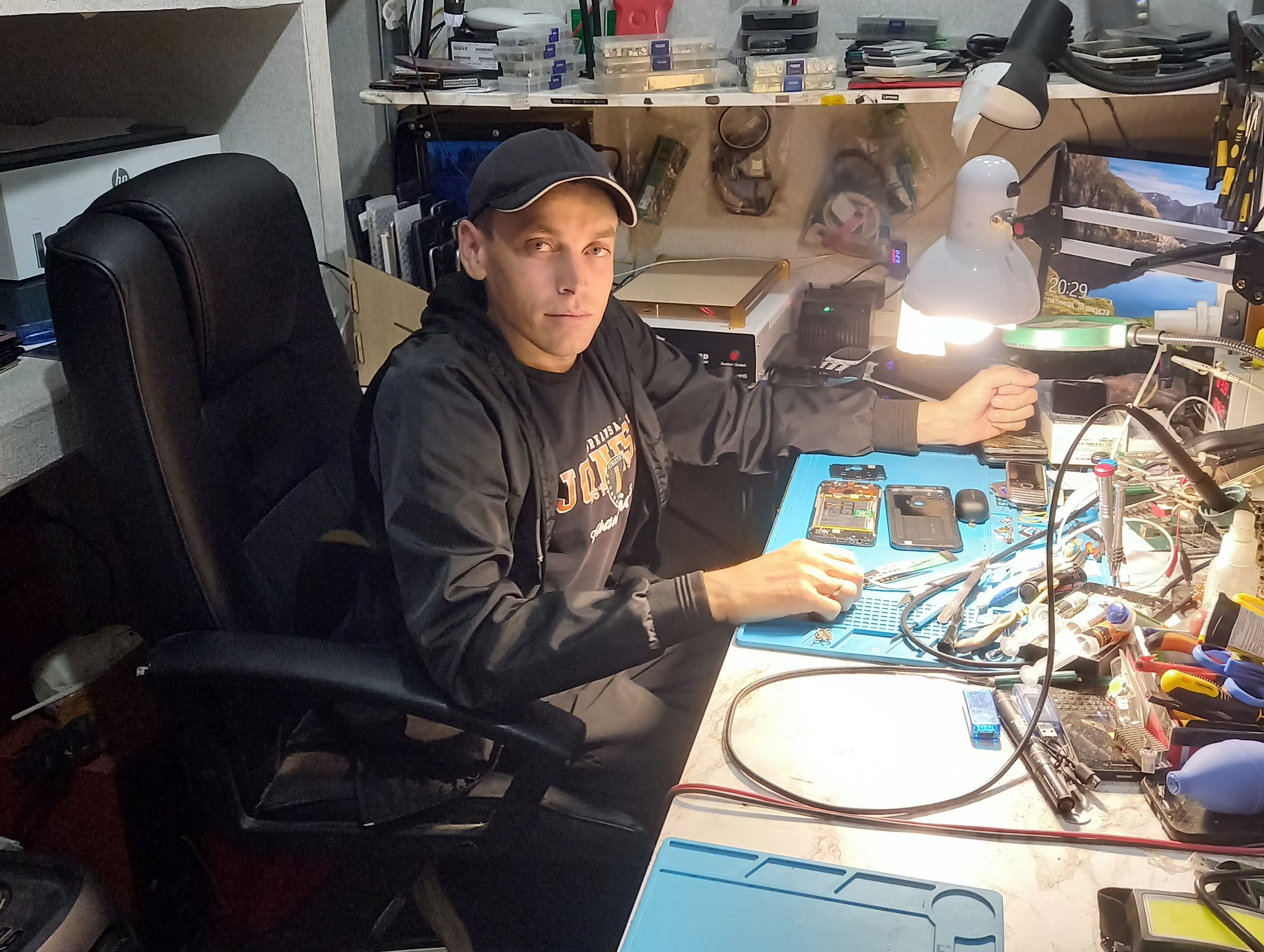 A man in a black shirt and cap sits at a desk where he assembles and disassembles a cell phone under a light.
