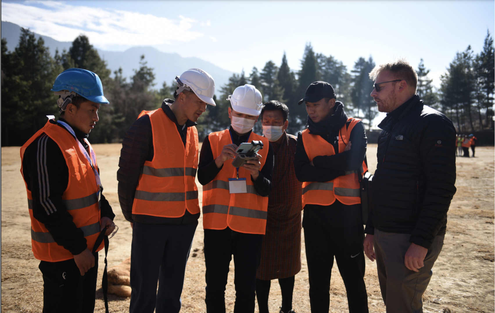 A group of five men, four of whom are in orange vests, gather around the remote controller of a drone