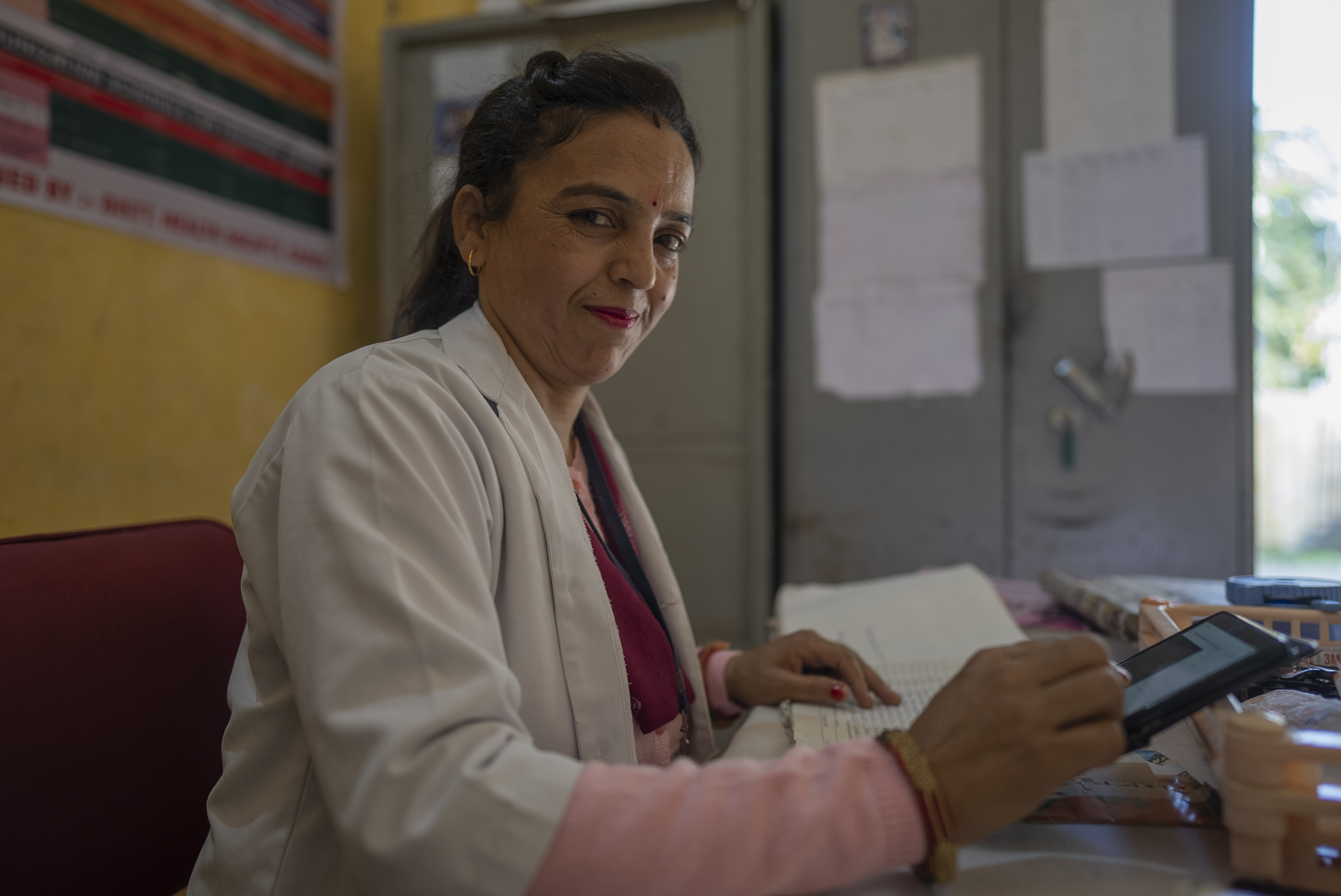 A woman in a white doctor's coat looks into the camera as she scrolls through a digital tablet