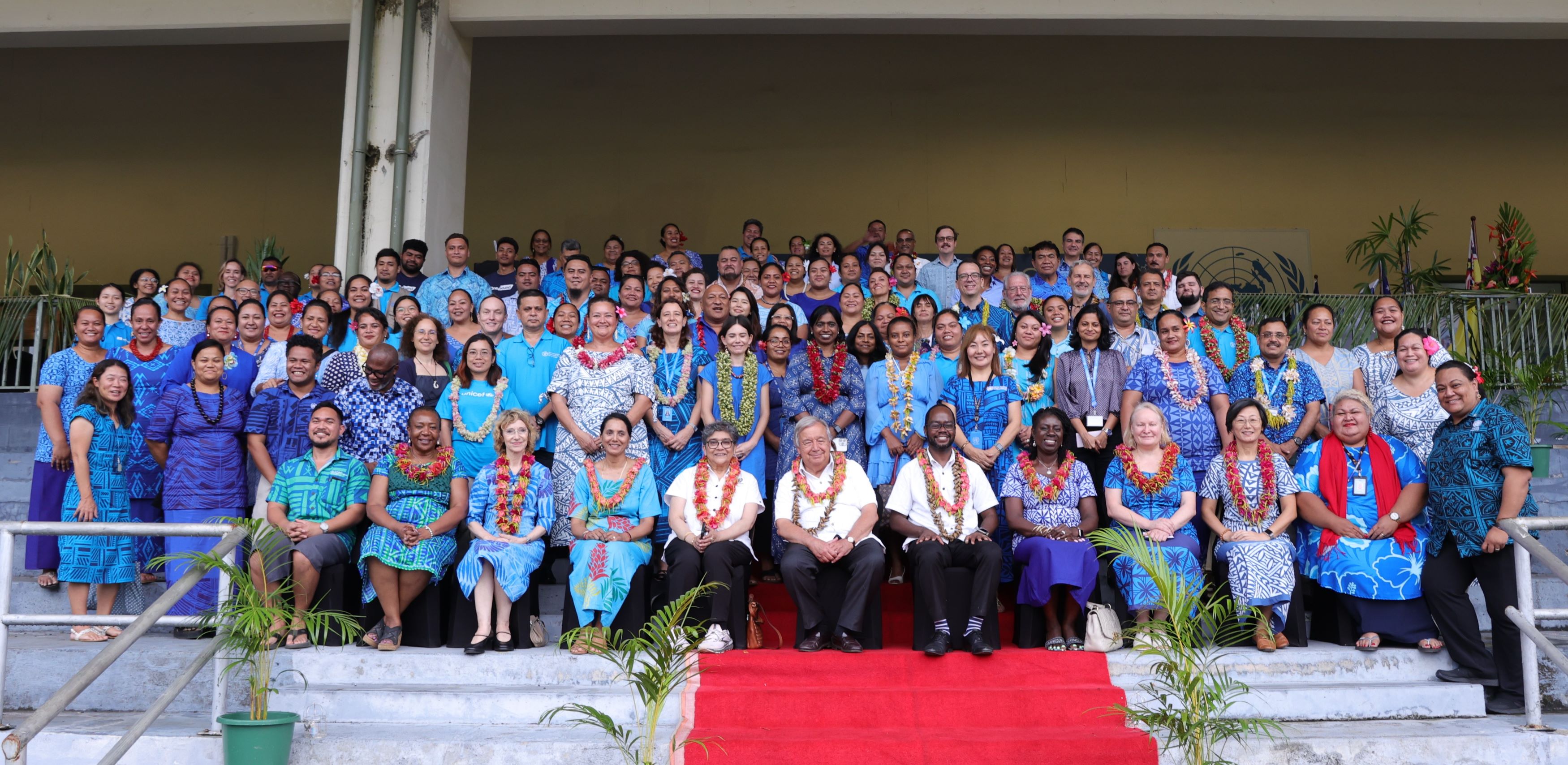 a group of people under a tent in an outdoor setting, they are all seated and wearing flower garlands and casual shirts