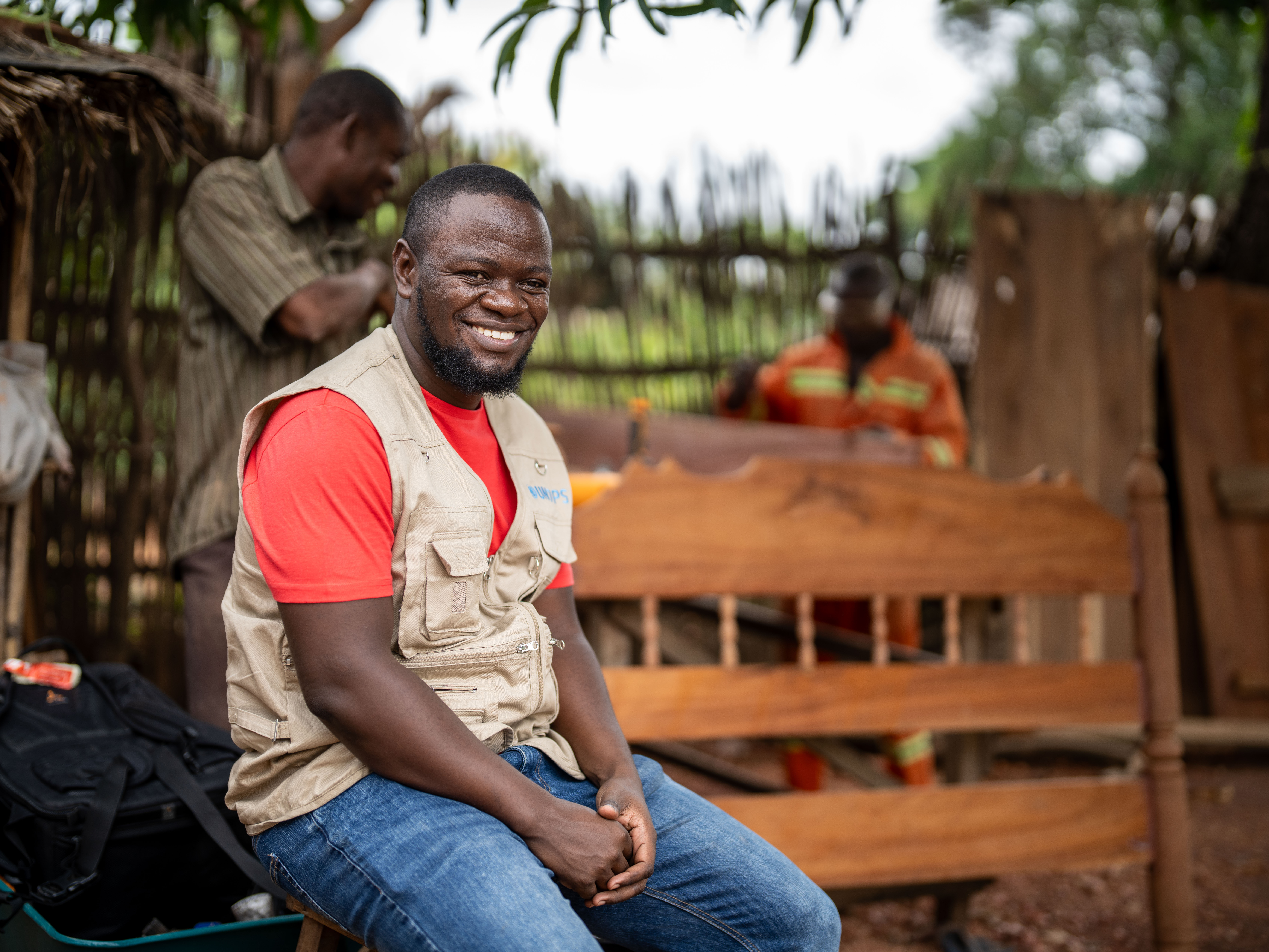 A man in a red shirt and grey vest sits on a wooden bench and smiles
