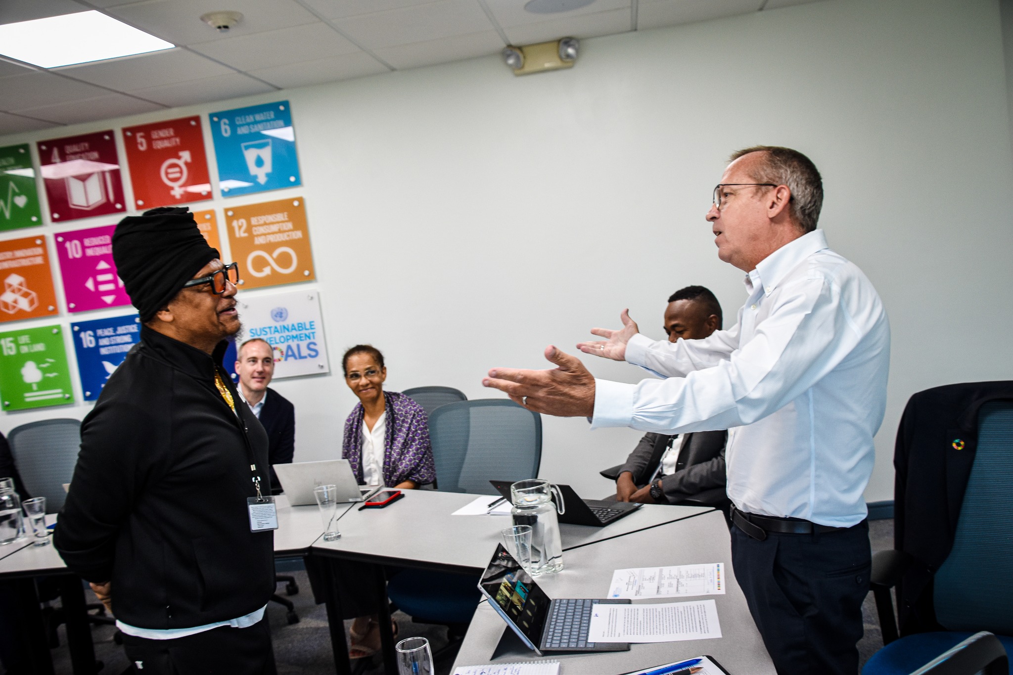 A man in a white shirt gesticulates as he speaks to another man in a dark shirt in a meeting room.