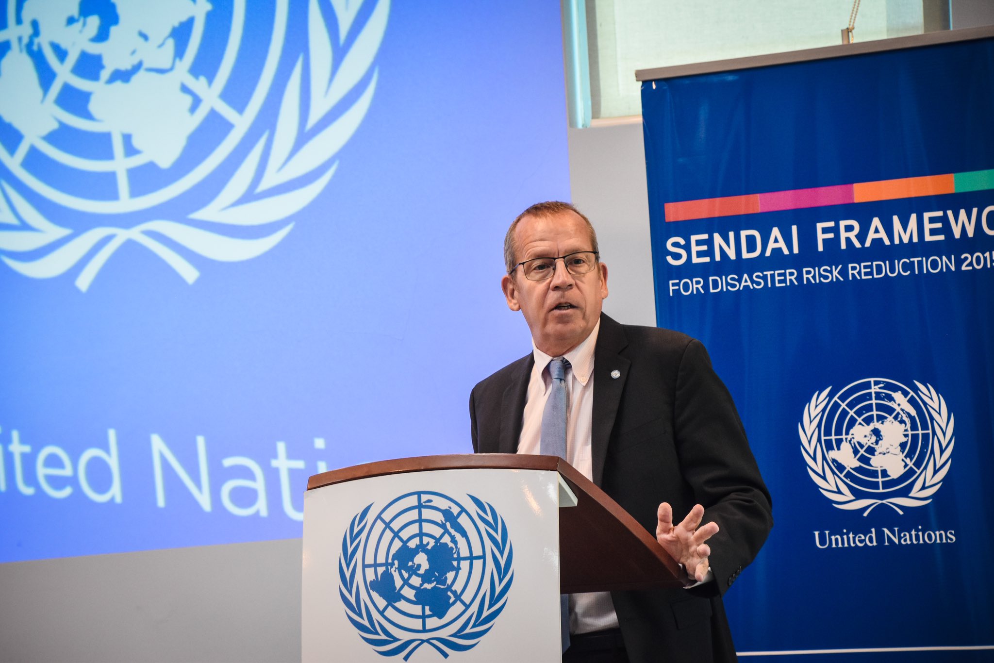 A man in a black suit speaks into a microphone at a podium against a blue background with the UN