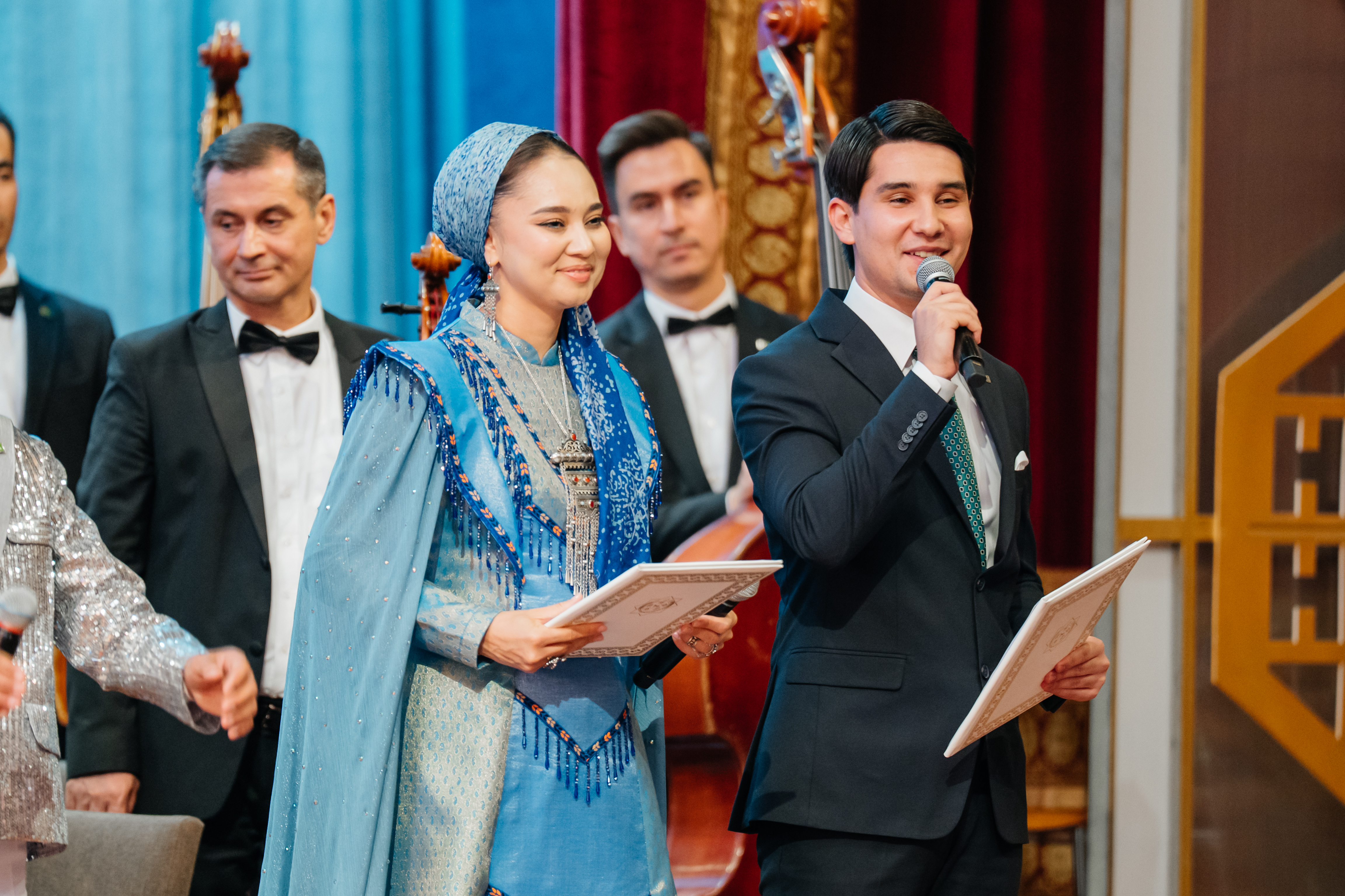 A man and woman in the foreground, with several people in the background, on stage at a concert in Turkmenistan.
