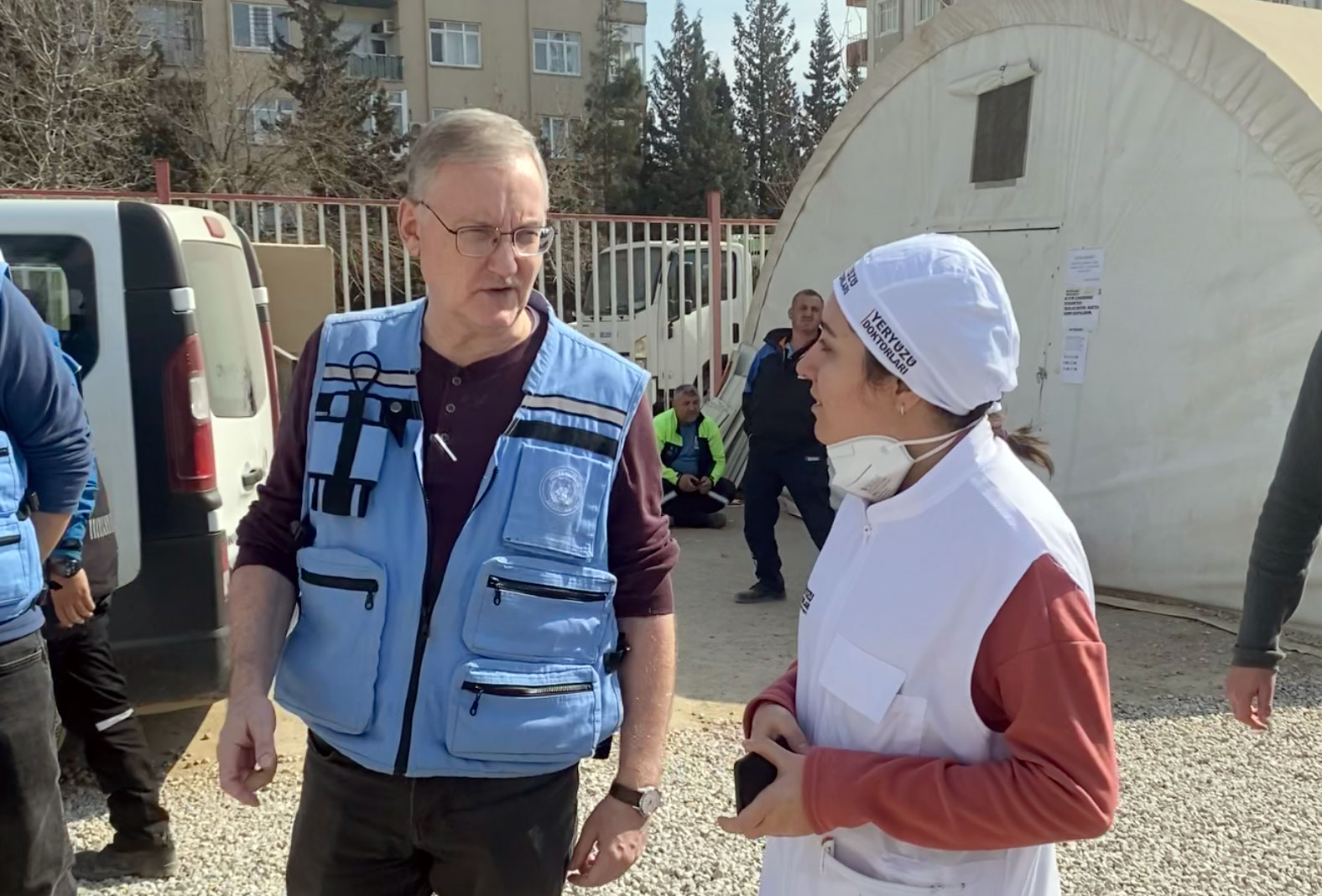 A man in blue humanitarian vest speaks to a women by a tent 