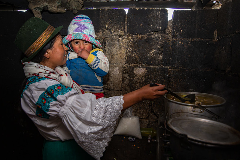 woman stirs a pot of soup while holding her young child 