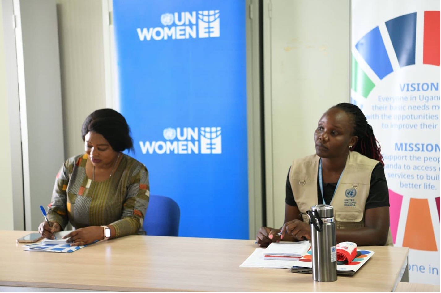 Two women sit at a table with a UN Women banner behind them 