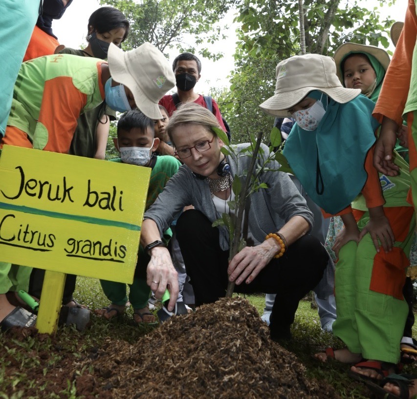 woman kneels down with children to plant tree 
