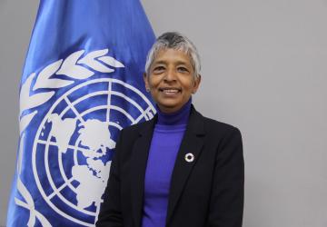 woman in dark suit standing in front of a UN flag 