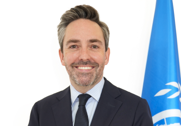 man in dark suit stands next to UN flag against white background