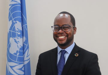man in dark suit and blue ties stands in front of UN flag