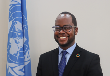 man in dark suit and blue ties stands in front of UN flag 