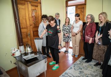 group of people gather in a circle in someone's home to watch a woman use a renewable energy stove. 