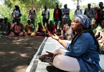 woman sits on the ground in blue tunic speaking to other women sitting in a circle 