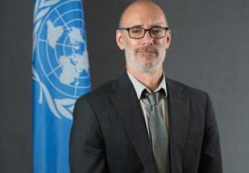 Man in a black suit and glasses stands in front of a blue UN flag