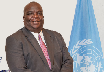 man in brown suit and pink tie stands in front of UN flag