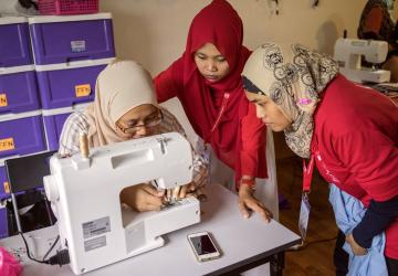 three women sit around a sewing machine wearing headscarves.