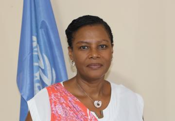 woman in white top and pink scarf looks into the camera standing in front of UN flag