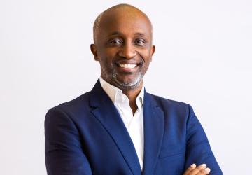 A man in a navy blue suit and a bright white collared shirt folds his arms and smiles for the camera.