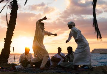 A group of men in white clothing dance together by a beachside as the sun sets in the background