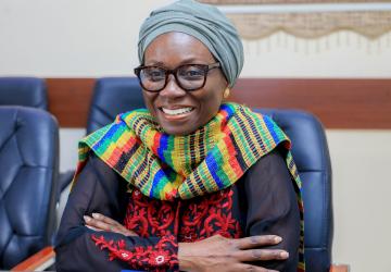 A woman in glasses and a colorful scarf and headwrap sits at a conference table, with hands folded, and smiles.