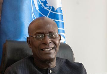 A man in a black buttoned-up blazer and glasses sits in front of a UN flag and smiles for the camera.
