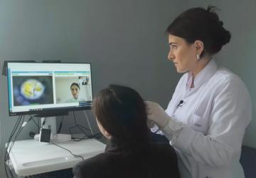 A doctor in a white coat diagnoses a woman in black clothing while another doctor is on a computer screen in front of them.