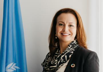 A woman in a black blazer and scarf stands next to a UN flag and smiles brightly.