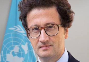 Joe Colombano wears a blue suite and red tie and stands in front of the UN flag