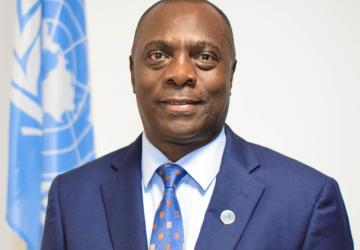A man in a blue suit and blue tie stands in front of a blue UN flag