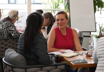 A woman in a red shirt talkng to another woman in a black shirt, there are other women in the background sitting on chairs and talking