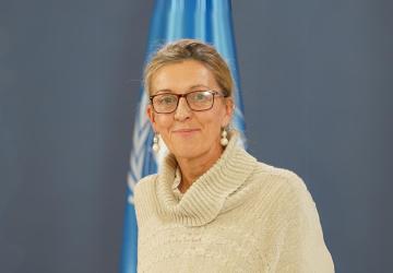 RC Françoise Jacob stands in front of a UN flag, she is wearing a cream coloured sweater