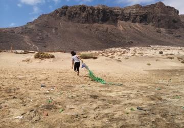 An individual dragging a large green net across a sandy area with sparse vegetation. In the background, there is a rugged mountain under a clear blue sky with few clouds. The ground is littered with various small pieces of debris.
