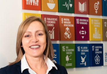 A woman wearing a white shirt and black jacket stands in front of colourful wallblocks that show the SDGs