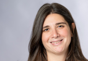 Kristèle Younès looks at the camera and smiles, she is standing in front of a grey background