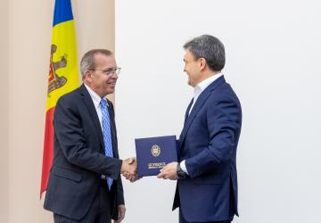 Two men in dark suits exchanging a document, shaking hands in front of a Moldovan flag. 