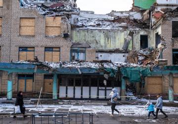 The image depicts a severely damaged multi-story building with its upper part largely destroyed, exposing the interior rooms. In the foreground, three people, including a child, walk past the building on snow-covered ground. 