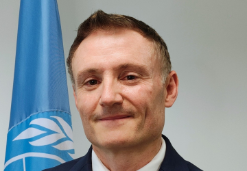 A man in a blue suit stands in front of a blue UN flag