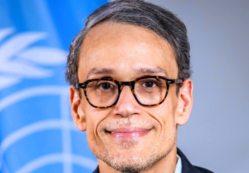 A man in a blue suit and black spectacles stands in front of a blue UN flag