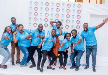 A group of nine young men and women in blue T shirts pose enthusiastically in front of a colourful background