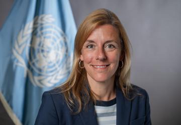 A woman with light brown hair and a dark suit stands in front of a grey background with a blue UN flag.