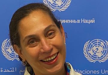 A woman wearing a white shirt looks at the camera and smiles with her head tilted, she is in front of a backdrop with the UN logo on it 