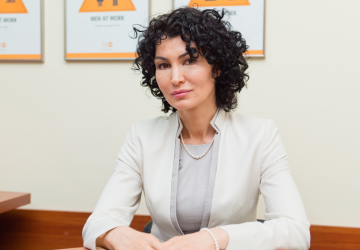 A woman wearing a white blazer and grey top looks at the camera. Behind her are posters saying men at work