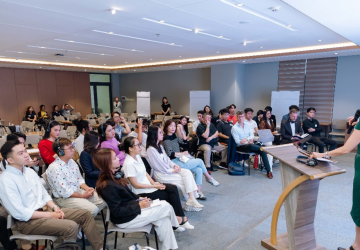 A woman is standing at the lectern and speaking to a group of young people in a conference room