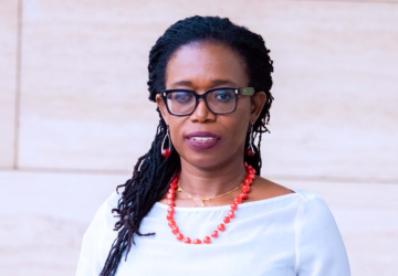 A woman looks at the camera wearing a white top and orange beaded necklace