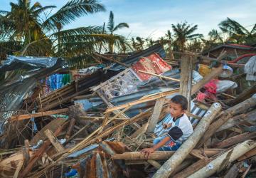 A collapsed house is in the background with a child seated on the rubble and crying 