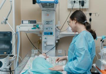 A woman is wearing a blue surgical cape and looking at her baby in an incubator. They are in a room with lots of medical equipment 