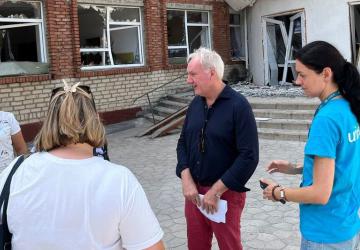 A man is wearing casual clothes and standing outdoors, he is surrounded by three women and stands in front of a partially destroyed building